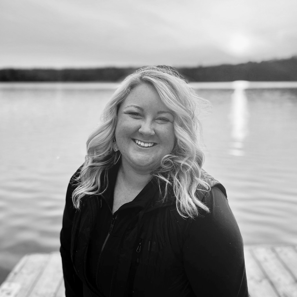 Headshot of Jocelyn hunt, smiling with curled blonde hair, against a landscape background of water and trees at sunset.